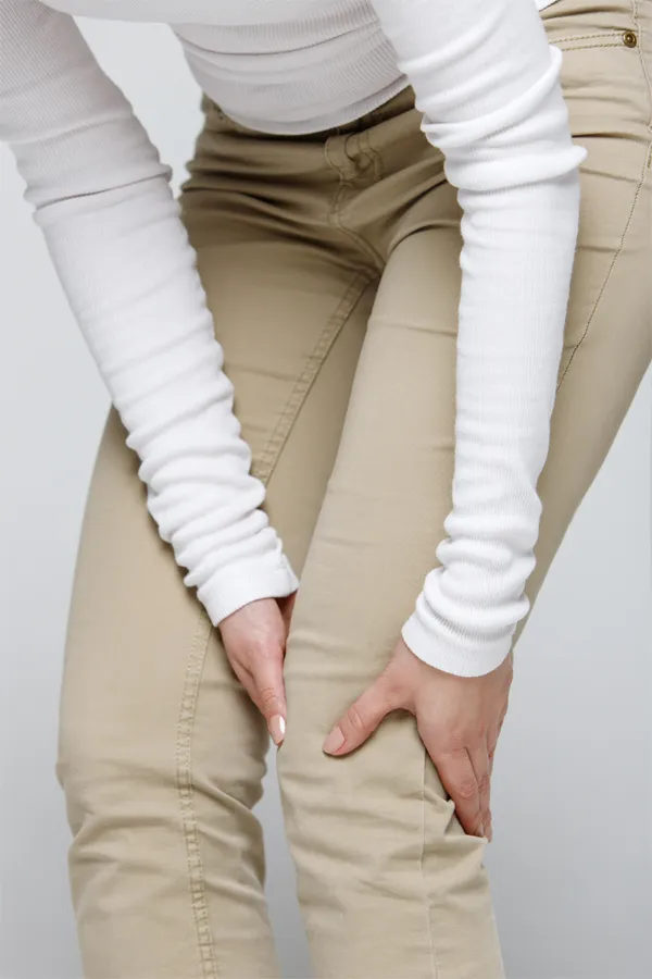 A woman wearing a white long-sleeve shirt and tan pants, clutching her knee, suffering from arthritis. Get arthritis treatment from Dr. David Lans in New Rochelle.