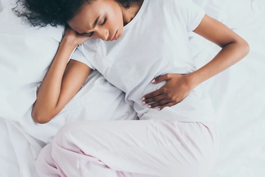 Crohns-Disease-Clinic A black woman in white pajamas lays on her side on white bed clutching her midsection. Get care for Crohn's Disease from Dr. David Lans in New Rochelle.