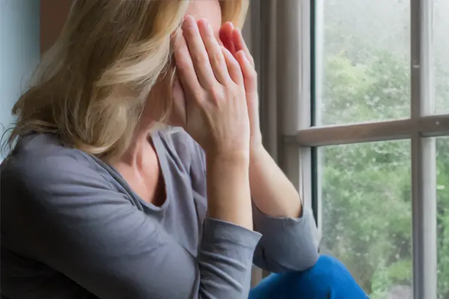 Graves-Disease-Clinic A blonde woman in a gray shirt sits in front of a window on a rainy day covering her eyes with her hands. Get treatment for Grave's Disease from Dr. David Lans in New Rochelle.