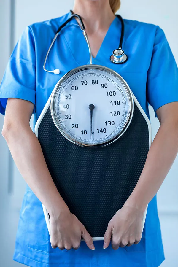 Medical-Weight-Loss-Treatment A nurse in blue scrubs with a stethoscope hanging around her neck holds a scale, representing weight loss treatments from Dr. David Lans in New Rochelle.