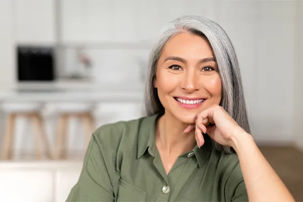 Menopause-Clinic An asian woman with long gray hair and a green shirt sitting and smiling on her couch, happy with her menopause care from [NAME} in New Rochelle.