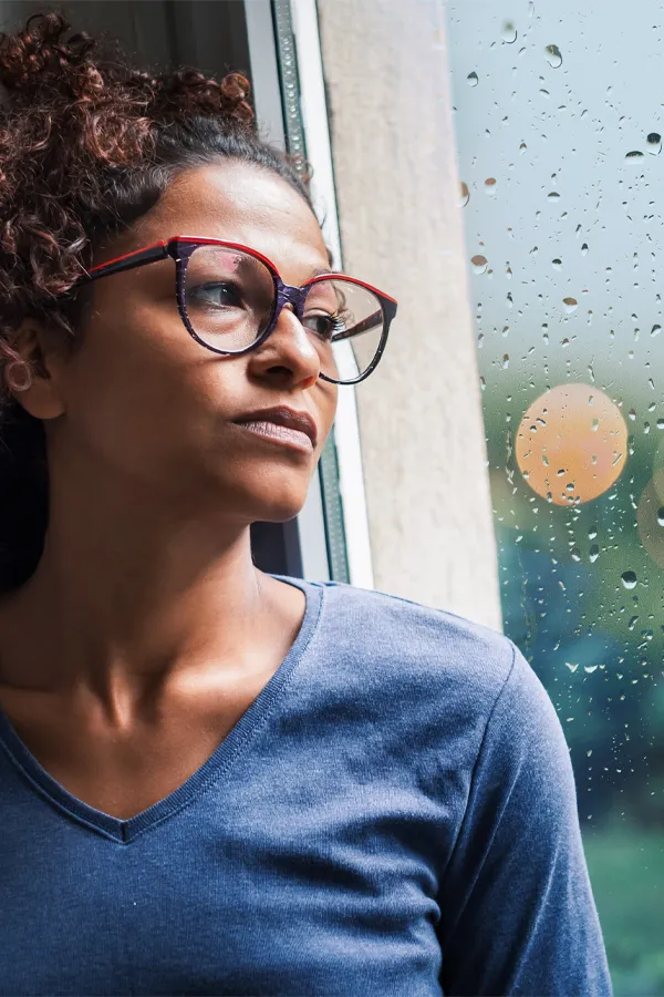 PMDD-Treatment A black woman with with a somber expression and glasses and a gray shirt stares out the window on a rainy day. Get treatment for Premenstrual Dysphoric Disorder from Dr. David Lans in New Rochelle.