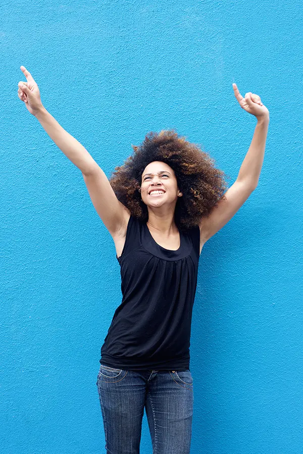 PMS-Treatment A woman in a dark blue tank top standing in front of a bright blue wall, raising her arms in celebration of relief from PMS from Dr. David Lans in New Rochelle.