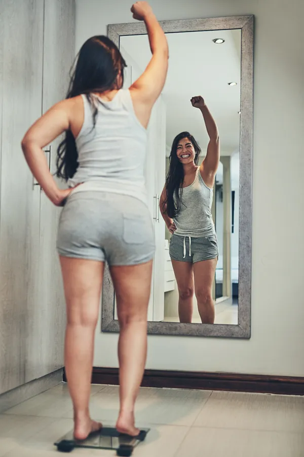 Tirzepatide-Treatment A woman in workout clothes stands on a scale looking in the mirror and pumping her hands in celebration of her successful Tirzepatide treatment from Dr. David Lans in New Rochelle.